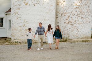 Family of four holding hands and walking on a gravel path in front of a weathered white brick silo during a casual outdoor photo session.