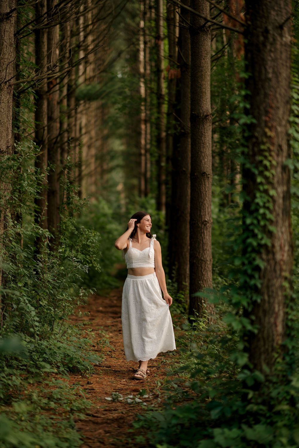 Woman in white crop top and skirt walking on a forest path surrounded by tall trees and lush greenery.