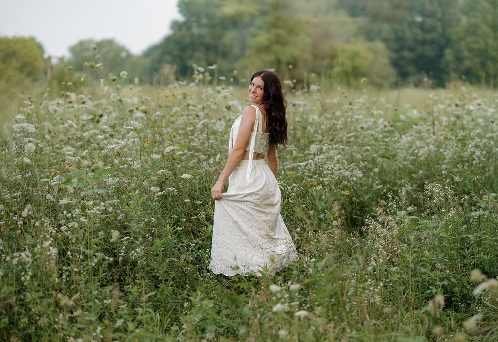 A woman in a white dress walks through a lush wildflower meadow, glancing back over her shoulder.