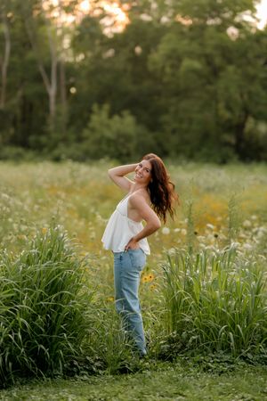 Young woman in white top and jeans posing in a wildflower meadow at golden hour with lush green trees behind her.
