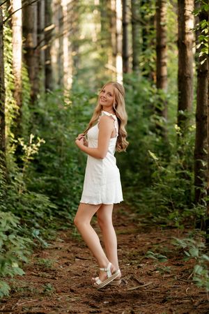 Young blonde woman in white dress posing on a forest path surrounded by tall trees and lush green foliage.