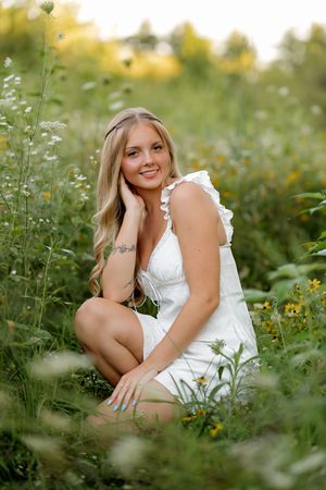 Young blonde woman in white dress sitting among wildflowers in a sunny green field, smiling warmly at camera.