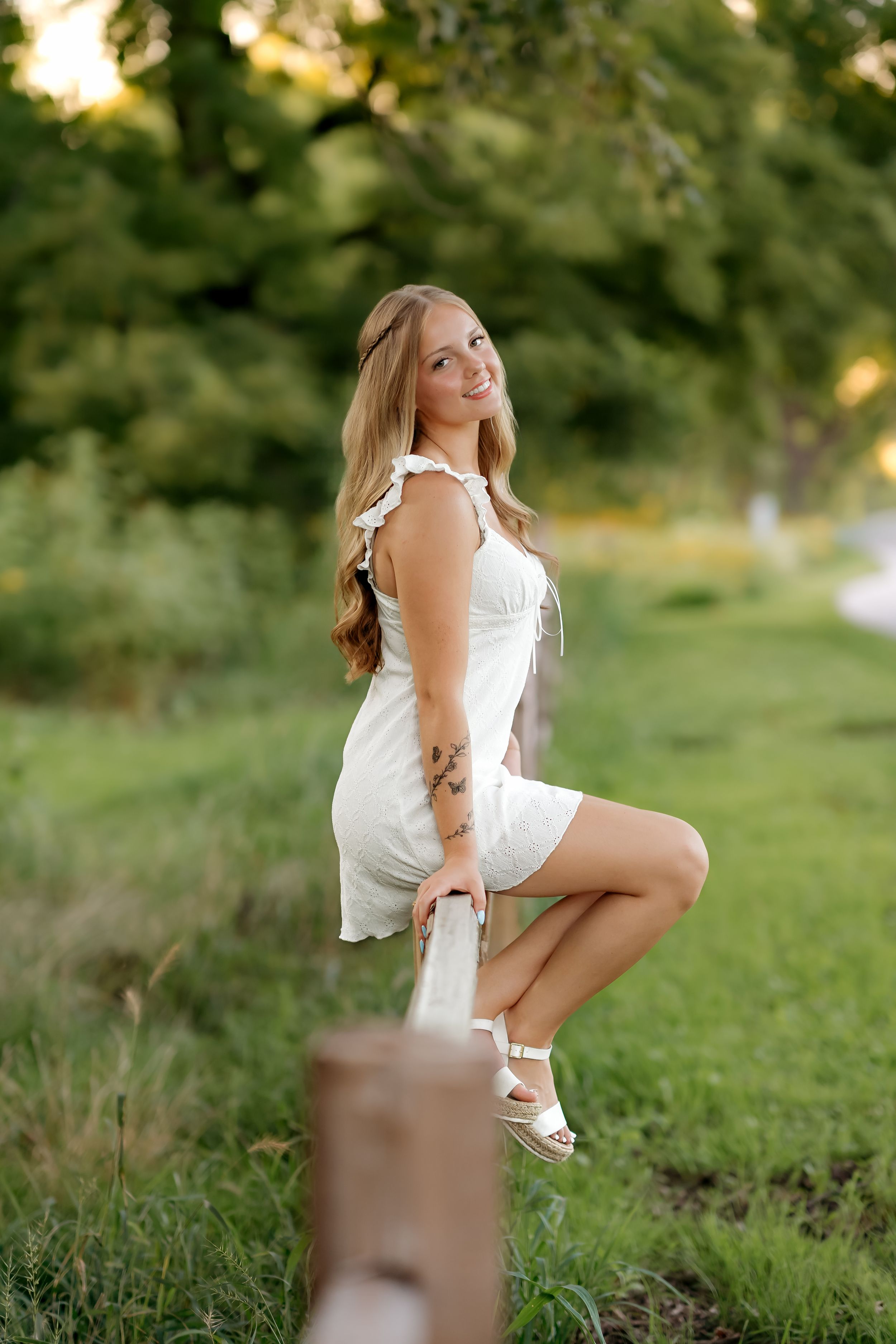 Young woman in white dress and sandals posing outdoors on a sunny day near a wooden fence with lush green trees.