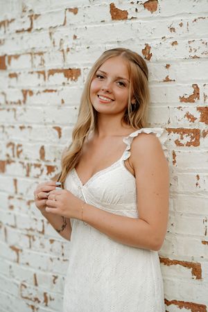 Smiling young woman in white lace dress leaning against rustic white brick wall with peeling paint.
