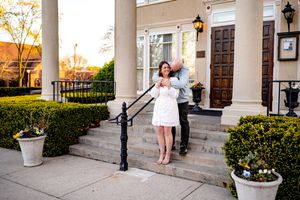 Someone in a white dress stands on the pillared entrance steps of an elegant building with ornate railings at sunset.