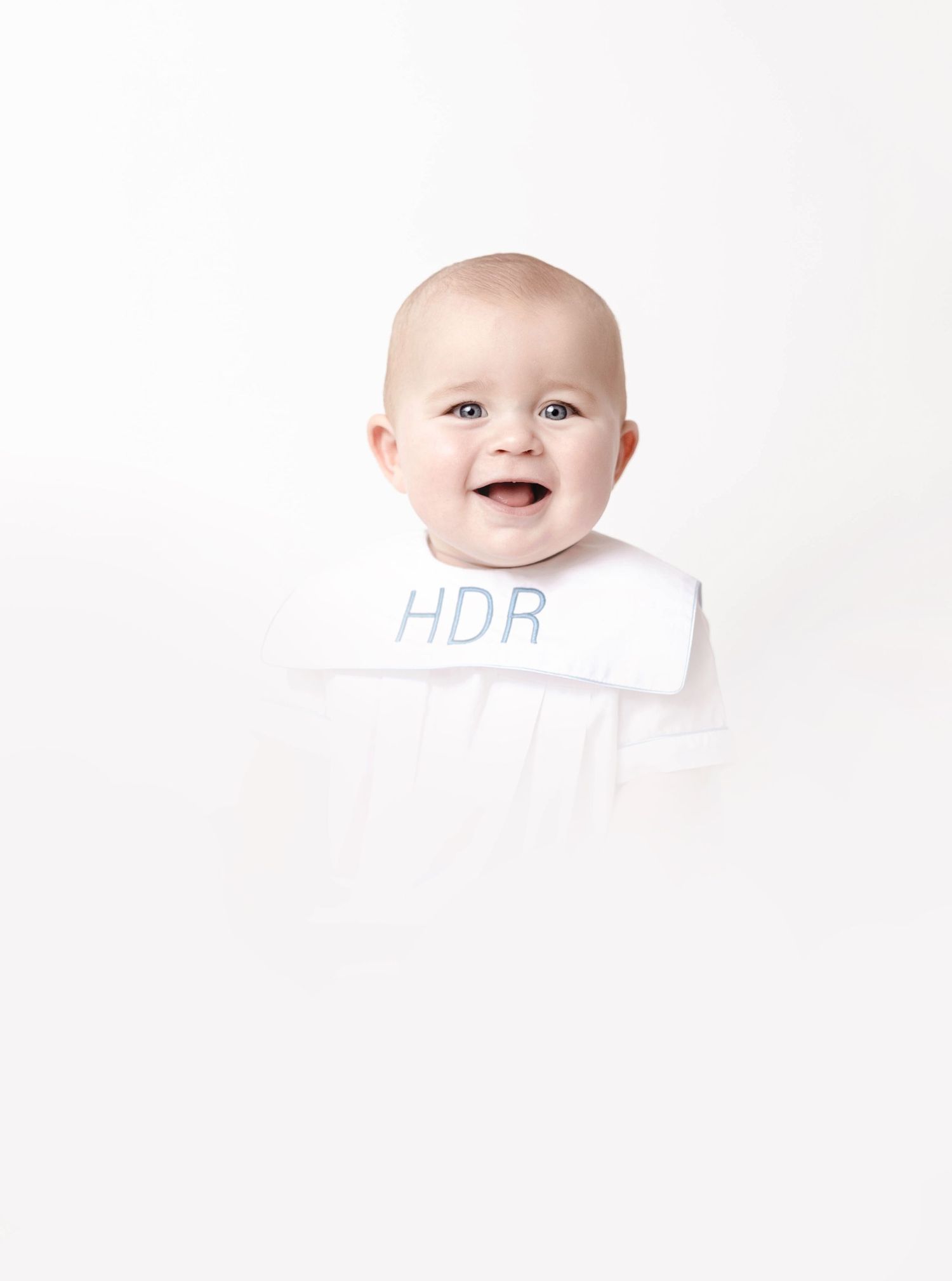 Smiling baby wearing white HDR monogrammed outfit against minimalist white backdrop.