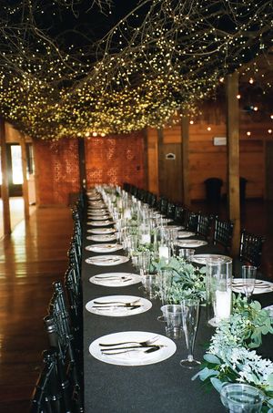 Elegant wedding reception table with black tablecloth and fairy lights overhead in rustic wooden venue.