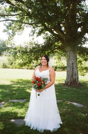 Beautiful summer wedding scene under a large tree with white gown and red floral bouquet on a sunny day.