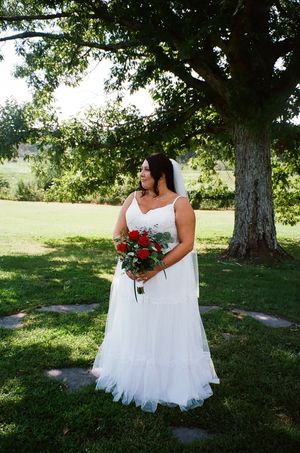 A bride in a flowing white dress holds a vibrant red bouquet while standing under a large tree in a sunlit garden.