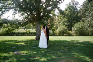 A couple stands together under a large tree on a sunny summer day in a lush green garden setting.
