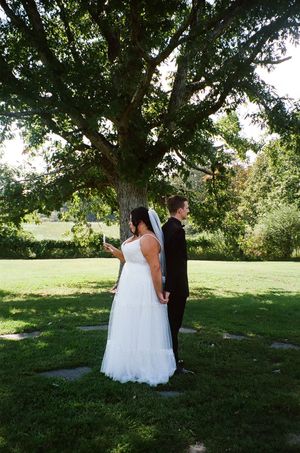 A couple in wedding attire stands beneath a large shady tree on a sunny summer day in a peaceful garden setting.