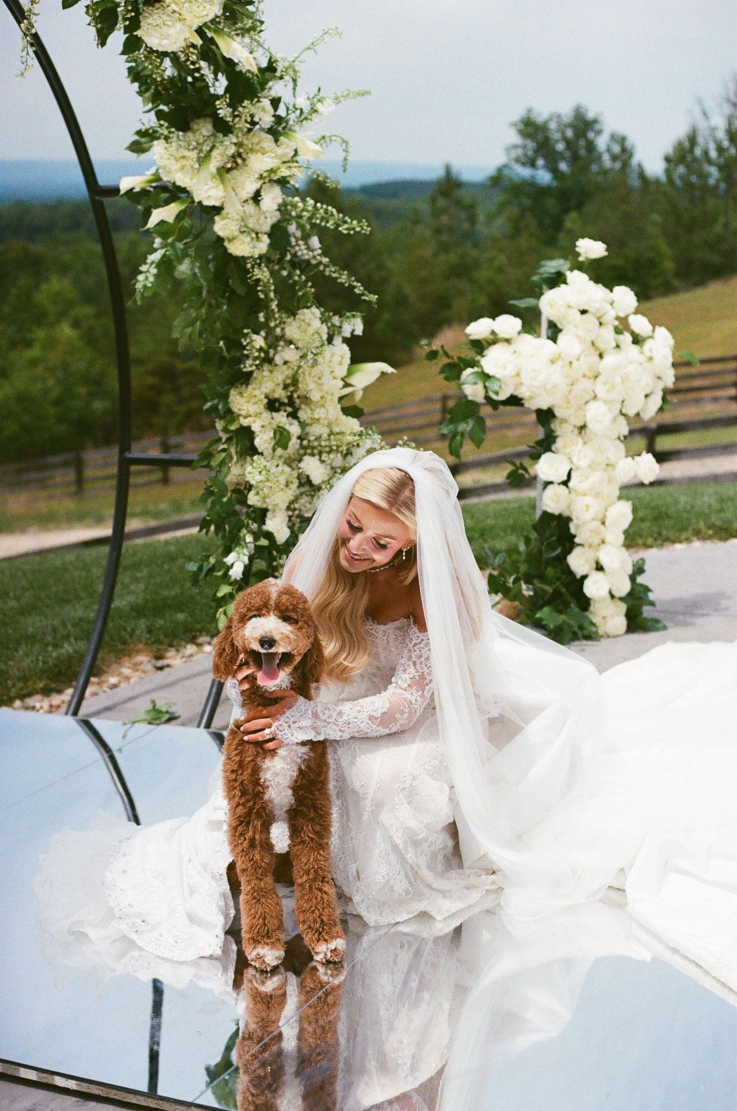 Bride in white lace gown and veil holding a fluffy golden doodle dog at an outdoor wedding ceremony with floral arch.
