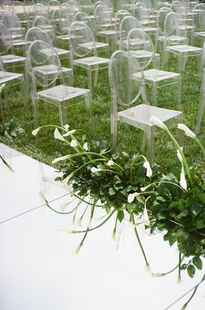 Clear ghost chairs arranged on green grass with white calla lily floral arrangements lining a white aisle runner at an outdoor wedding.