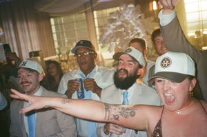 Group of friends celebrating at a wedding reception, wearing baseball caps, dancing and cheering with tattoos visible.
