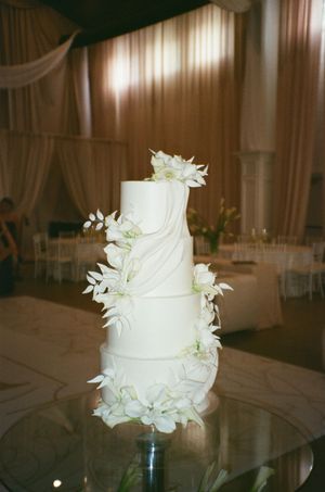 Elegant four-tier white wedding cake adorned with white flowers and draped fondant details on a glass stand in a ballroom.