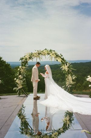 Bride and groom exchange vows under a floral arch on a mirrored platform with scenic green hills backdrop.