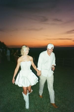 Couple in white outfits holding hands at sunset, woman in mini dress and cowboy boots, man in shirt and cap.