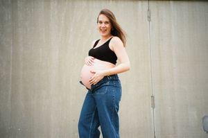 Maternity photo against concrete wall shows woman in casual attire holding her baby bump.