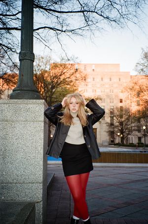 Person in red tights and black jacket poses playfully against stone pillar at sunset.