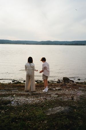 Two people stand together at water's edge on rocky shoreline during cloudy day.