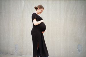 Expectant mother in black dress poses against light wall.