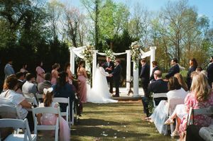 Outdoor wedding ceremony with white arch decorated with flowers and guests seated on white chairs.