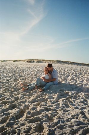 A couple embraces while sitting on pristine white sand beach with sand dunes and blue sky in background.