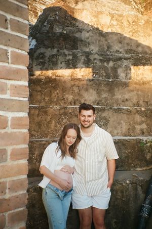 A couple poses against a rustic brick wall during a maternity photo shoot outdoors.