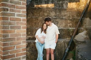 A couple stands against weathered brick wall during casual maternity photo session.