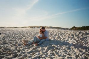 Couple sits close together on white sandy beach with dunes in background.
