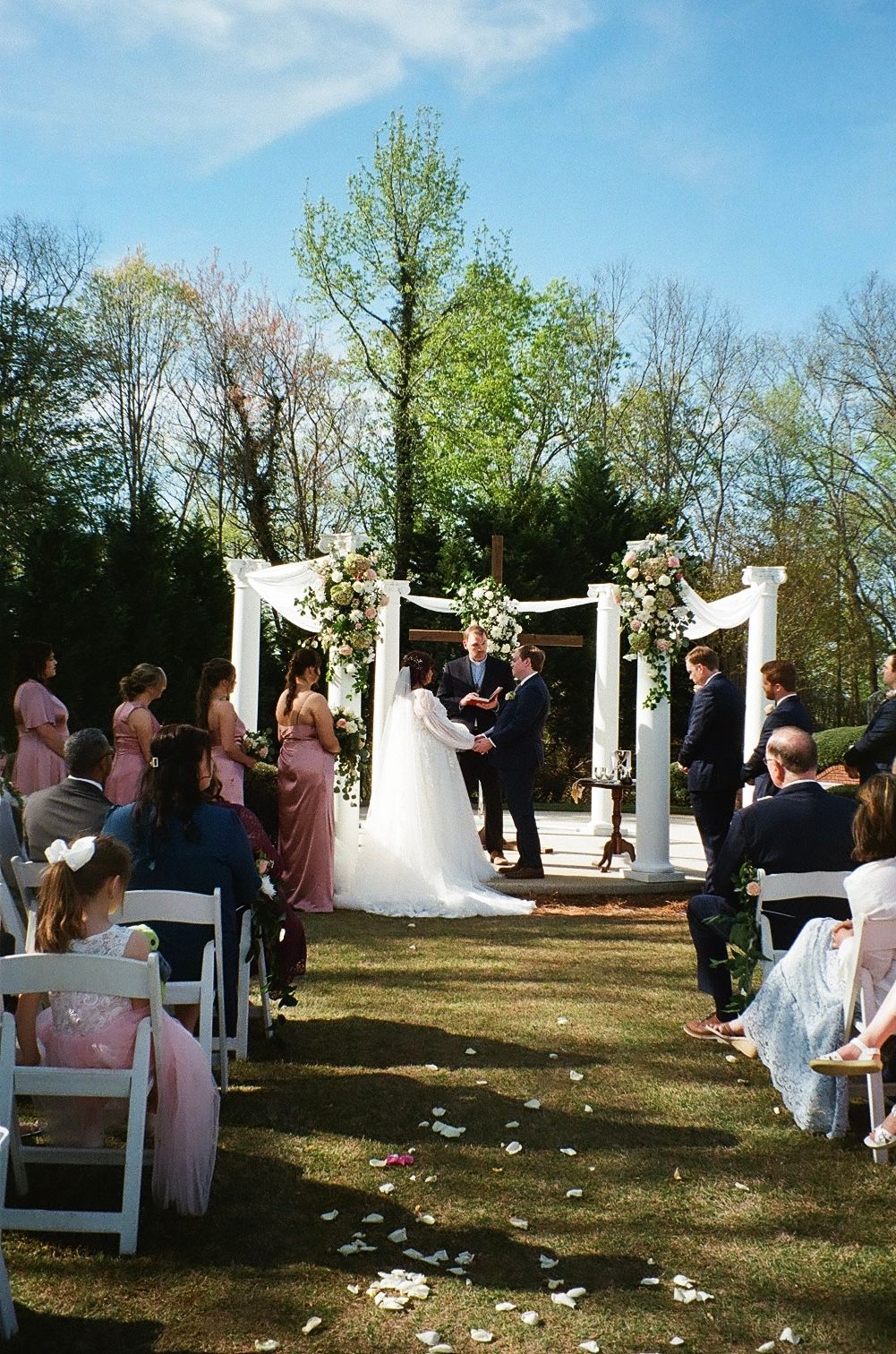 Outdoor wedding ceremony under a white arch decorated with flowers.