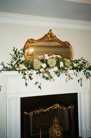 Ornate gold mirror above fireplace mantel decorated with white flowers and greenery.