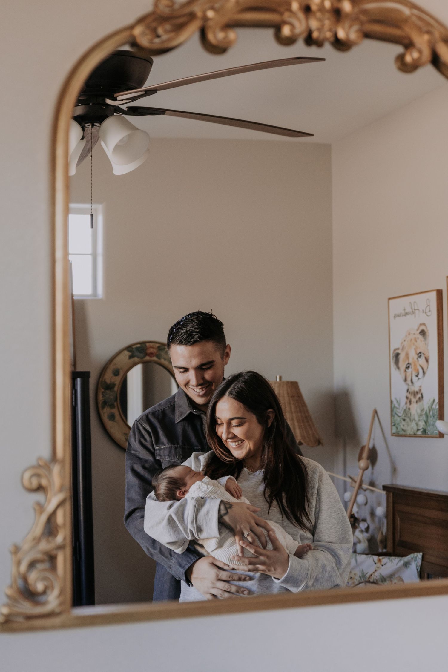 Happy couple smiling at newborn baby reflected in ornate gold-framed mirror.