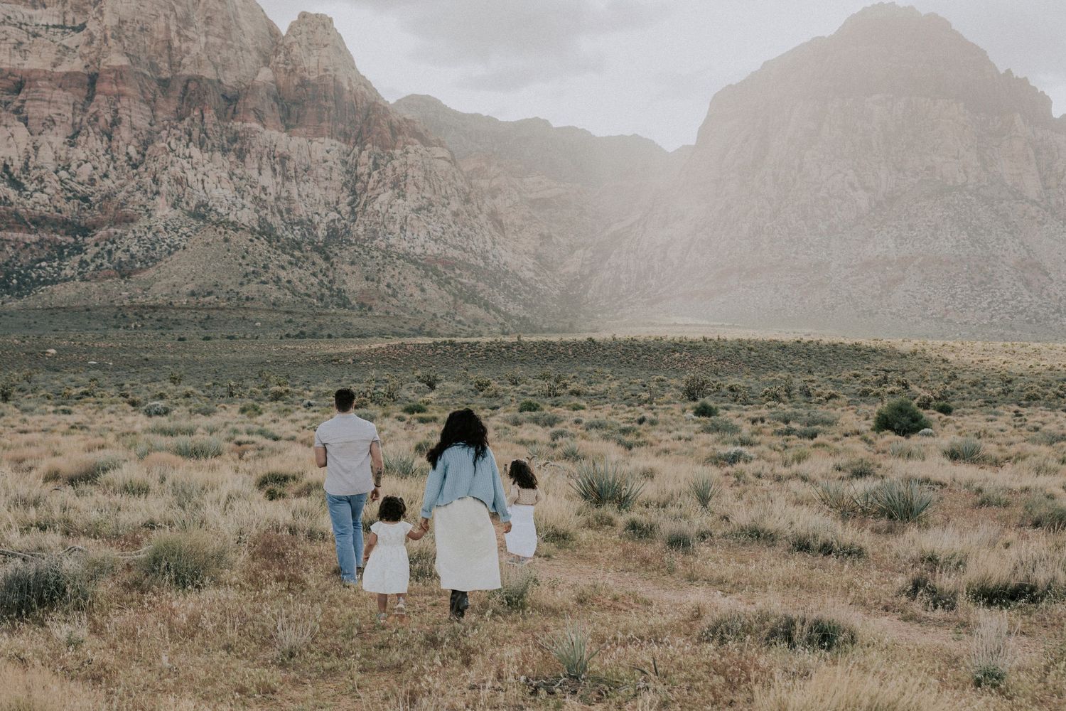 Family of four walking hand-in-hand toward Red Rock Canyon mountains in Nevada desert.