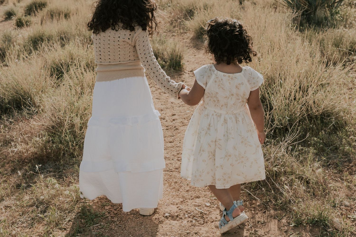 Mother and young daughter walking hand-in-hand through golden dry grass field.