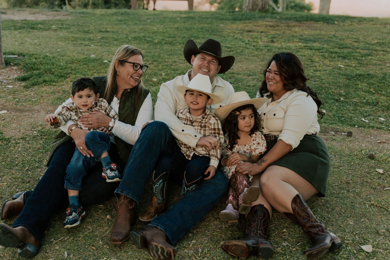 Casual family portrait of six people in cowboy hats sitting on grass in a sunny outdoor setting.