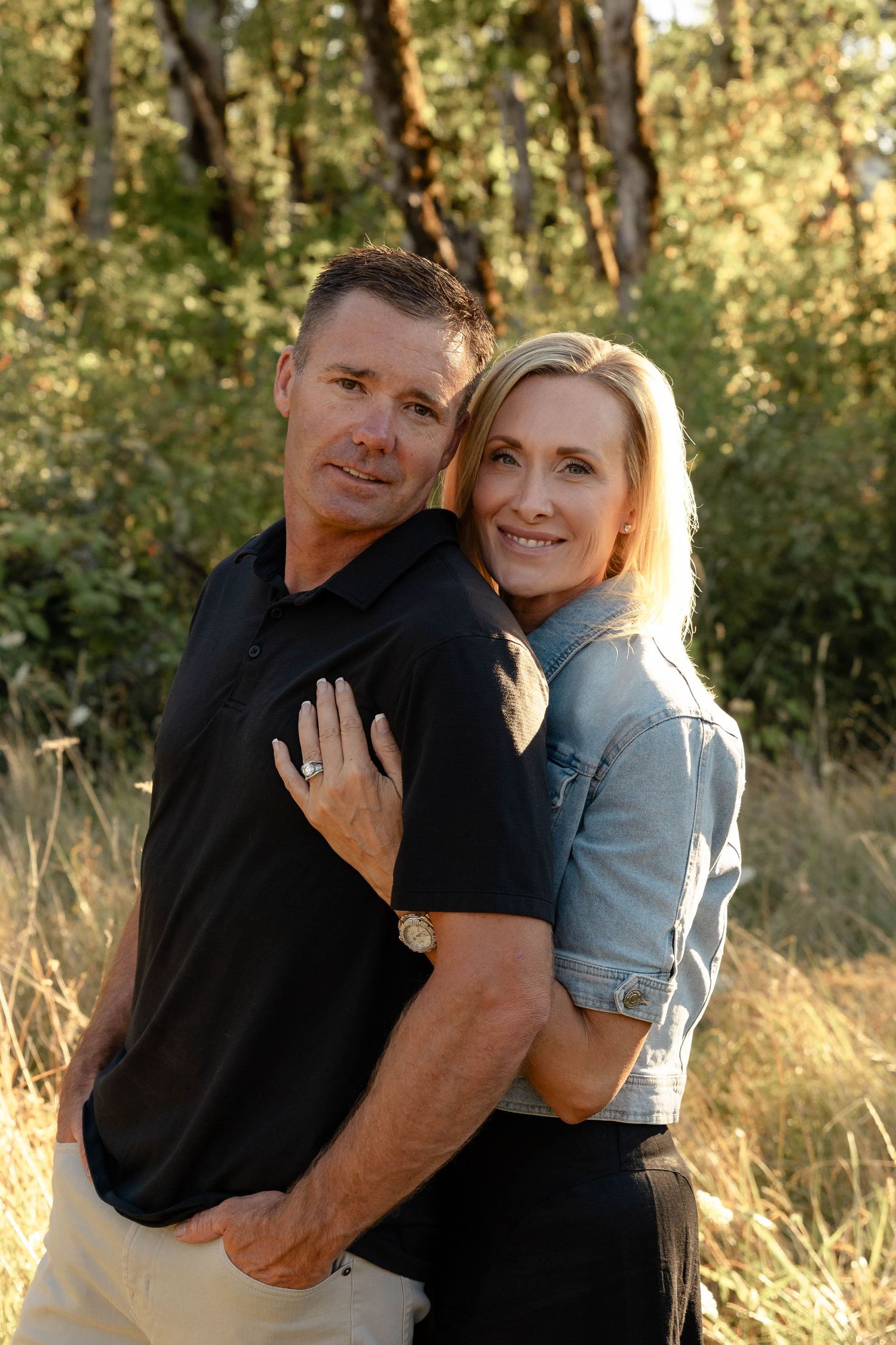 Smiling couple embracing outdoors in warm golden light with tall grass and trees behind them.