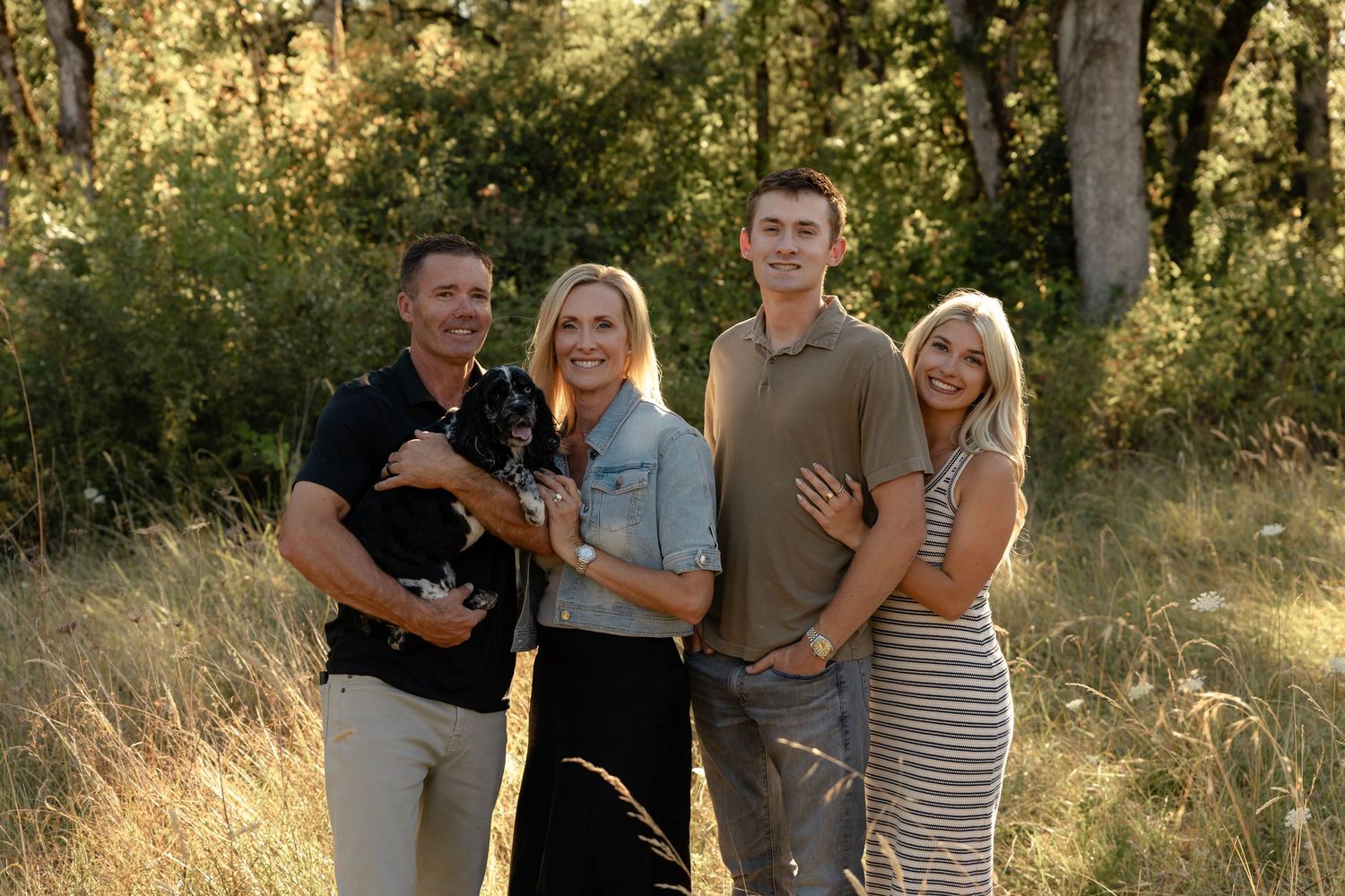Family of four posing together outdoors in golden sunlight with lush green trees in background.