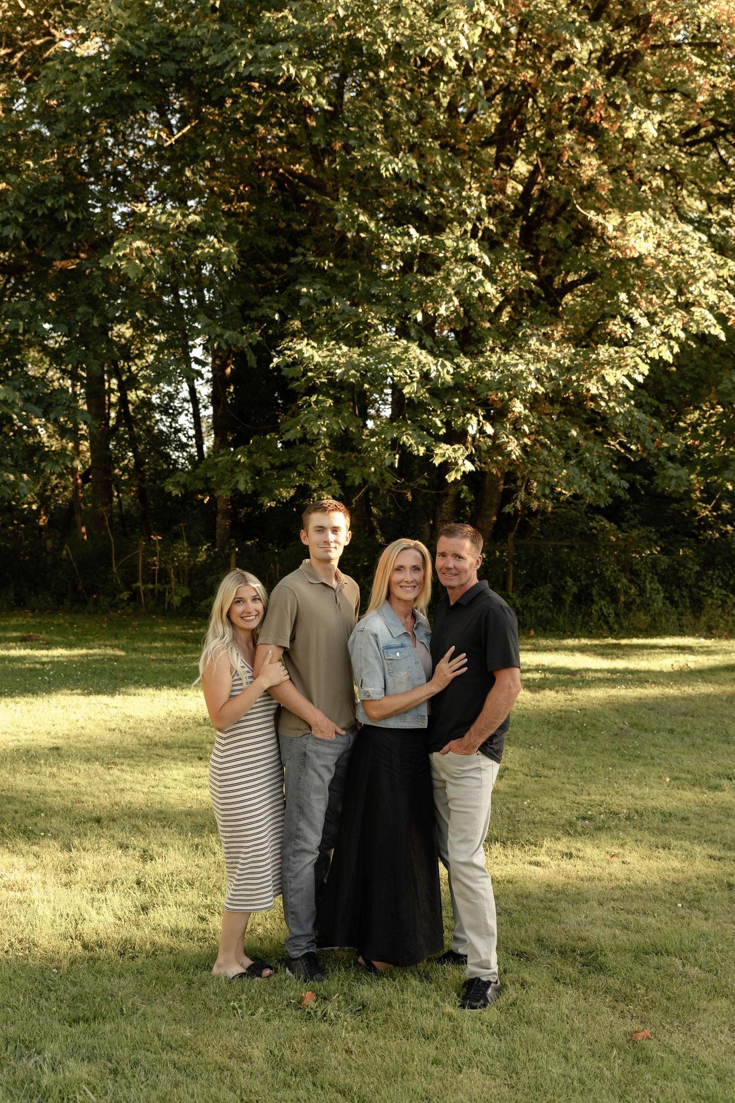 Happy family of four posing on green lawn surrounded by large leafy trees in warm sunlight.