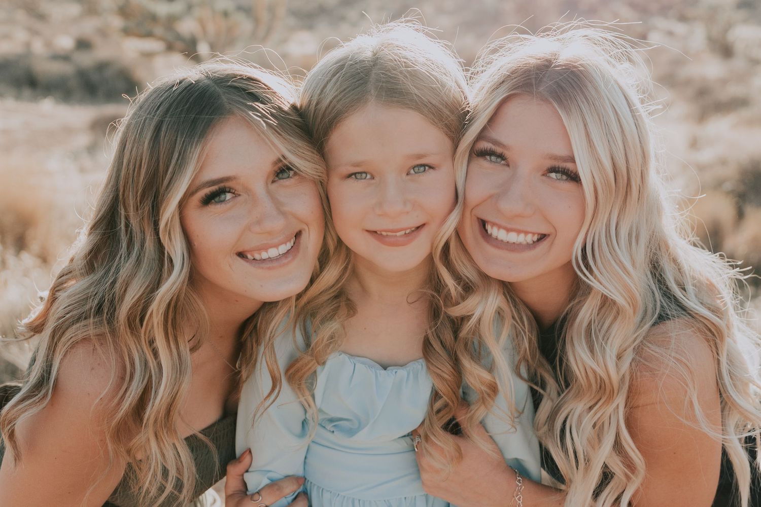 Three smiling girls with blonde hair posing together outdoors in desert setting.