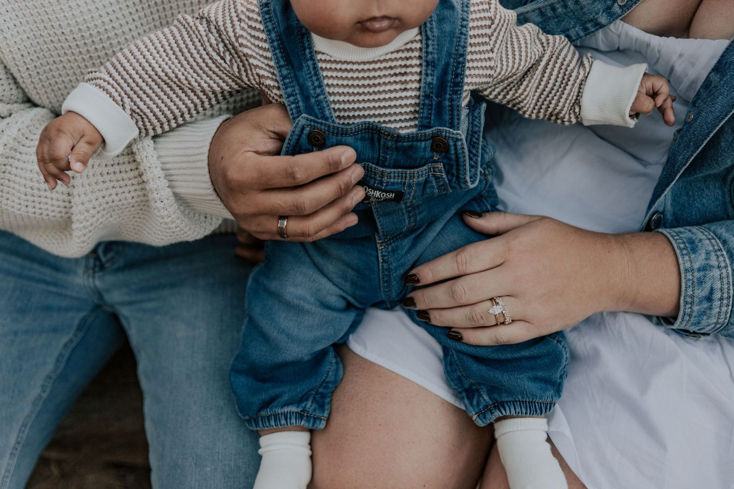 Close-up of parents holding baby dressed in denim overalls between them.