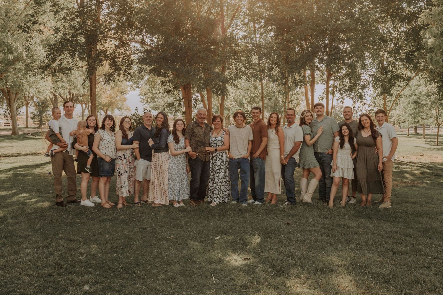 Large extended family group photo outdoors in a park with tall trees and golden sunlight.
