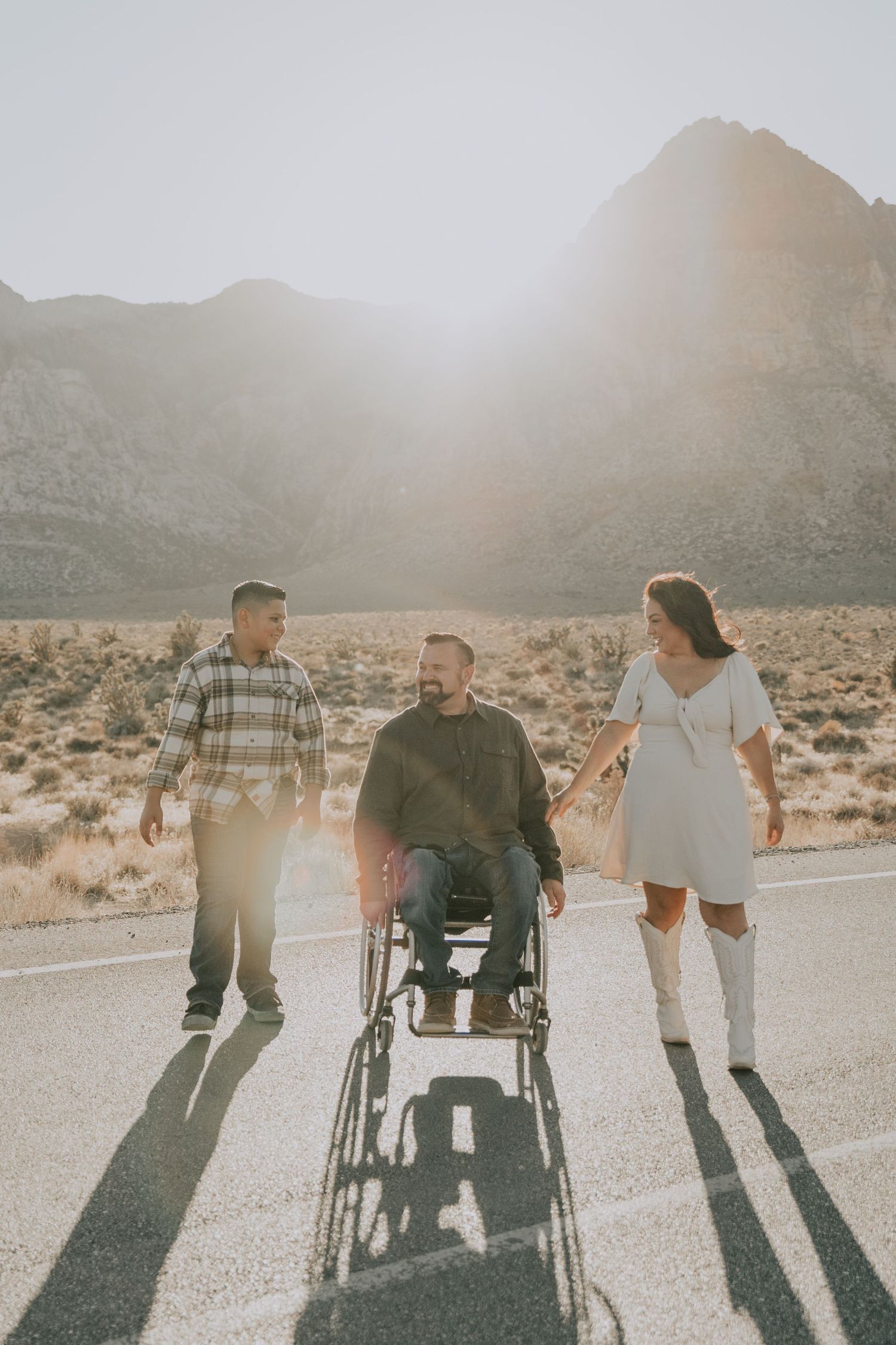 Man in wheelchair with two companions walking on desert road with mountain backdrop.