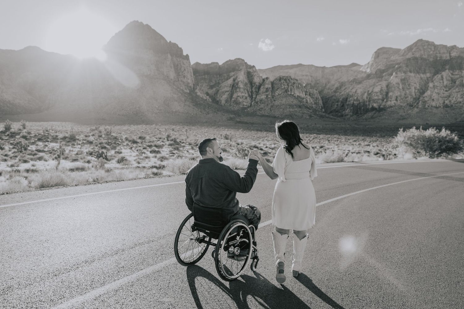 Black and white photo of couple on desert road, man in wheelchair holding woman's hand.