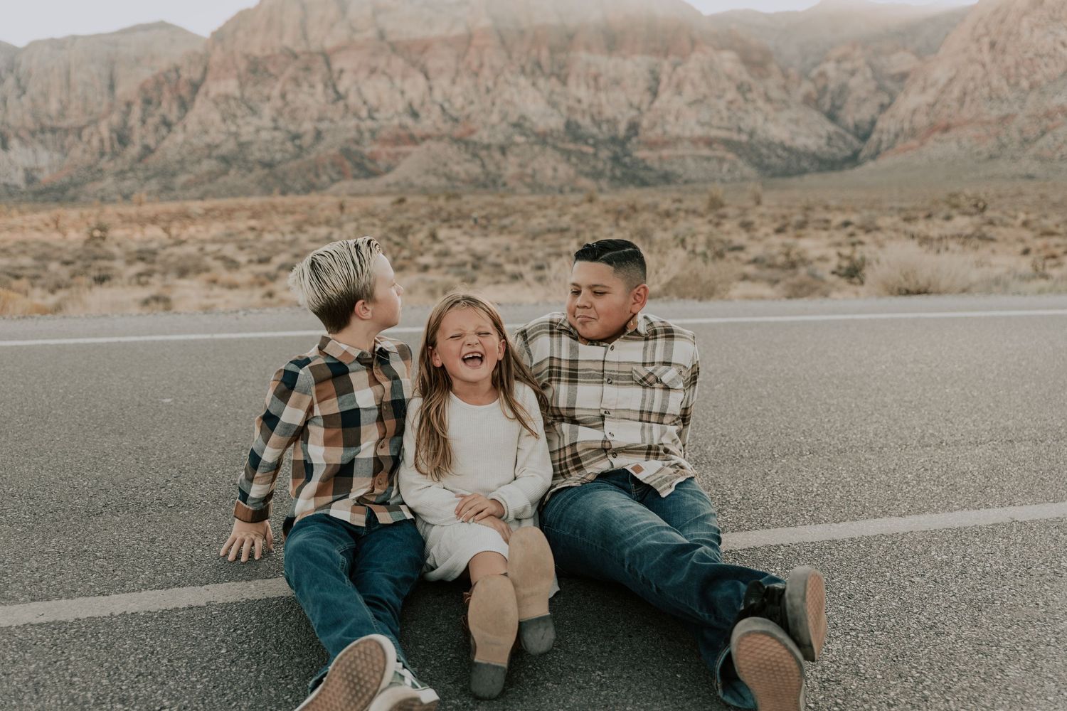 Three children laughing and sitting together on road with desert mountain scenery.