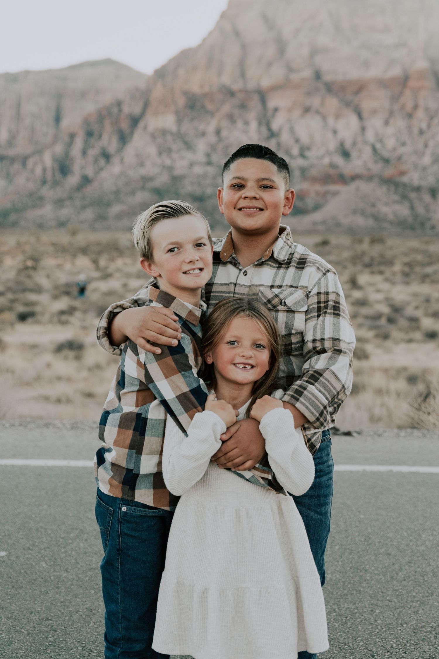 Three siblings hugging and smiling with red rock mountain backdrop outdoors.