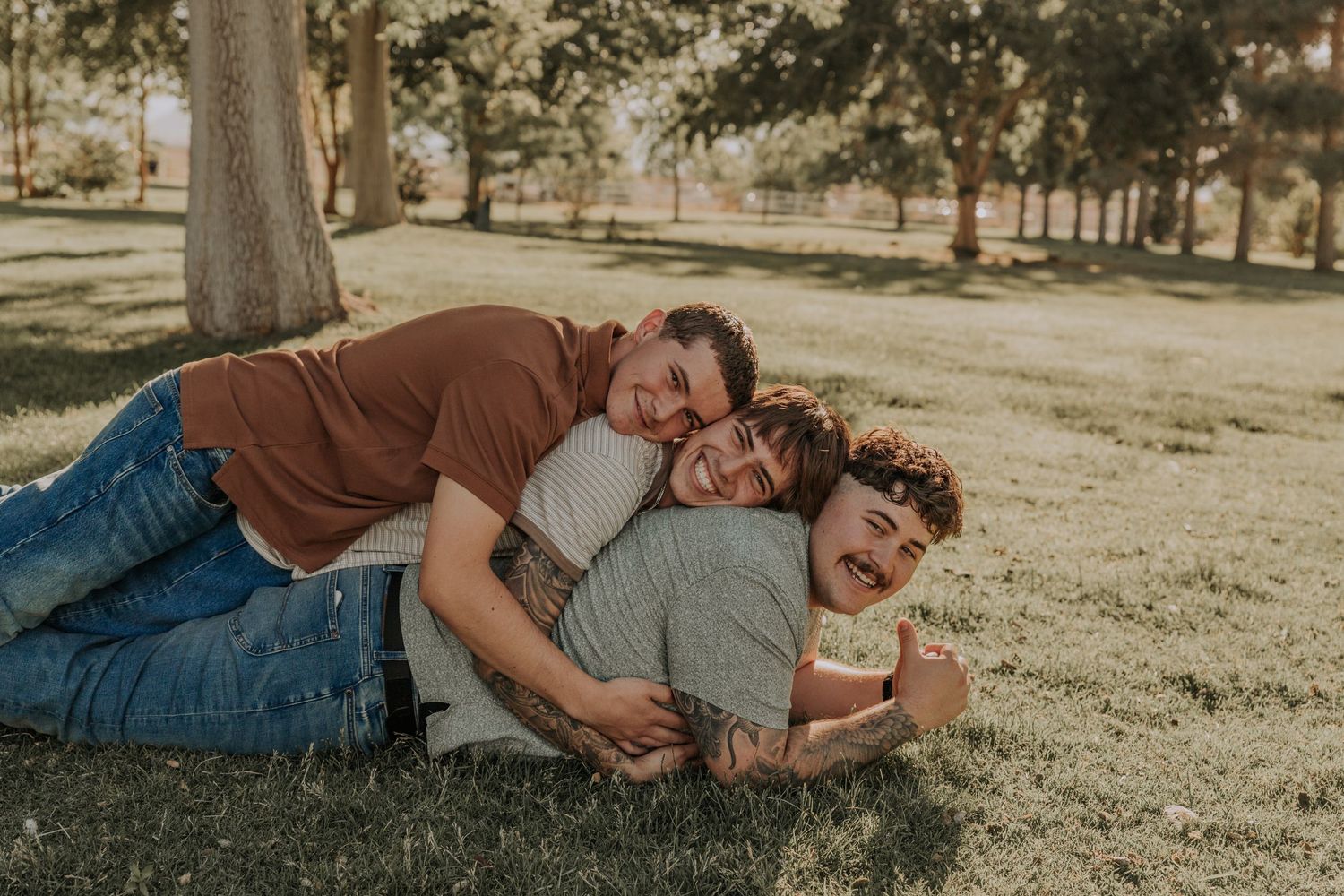 Three young men playfully piling on each other and laughing while lying on green grass in a park.