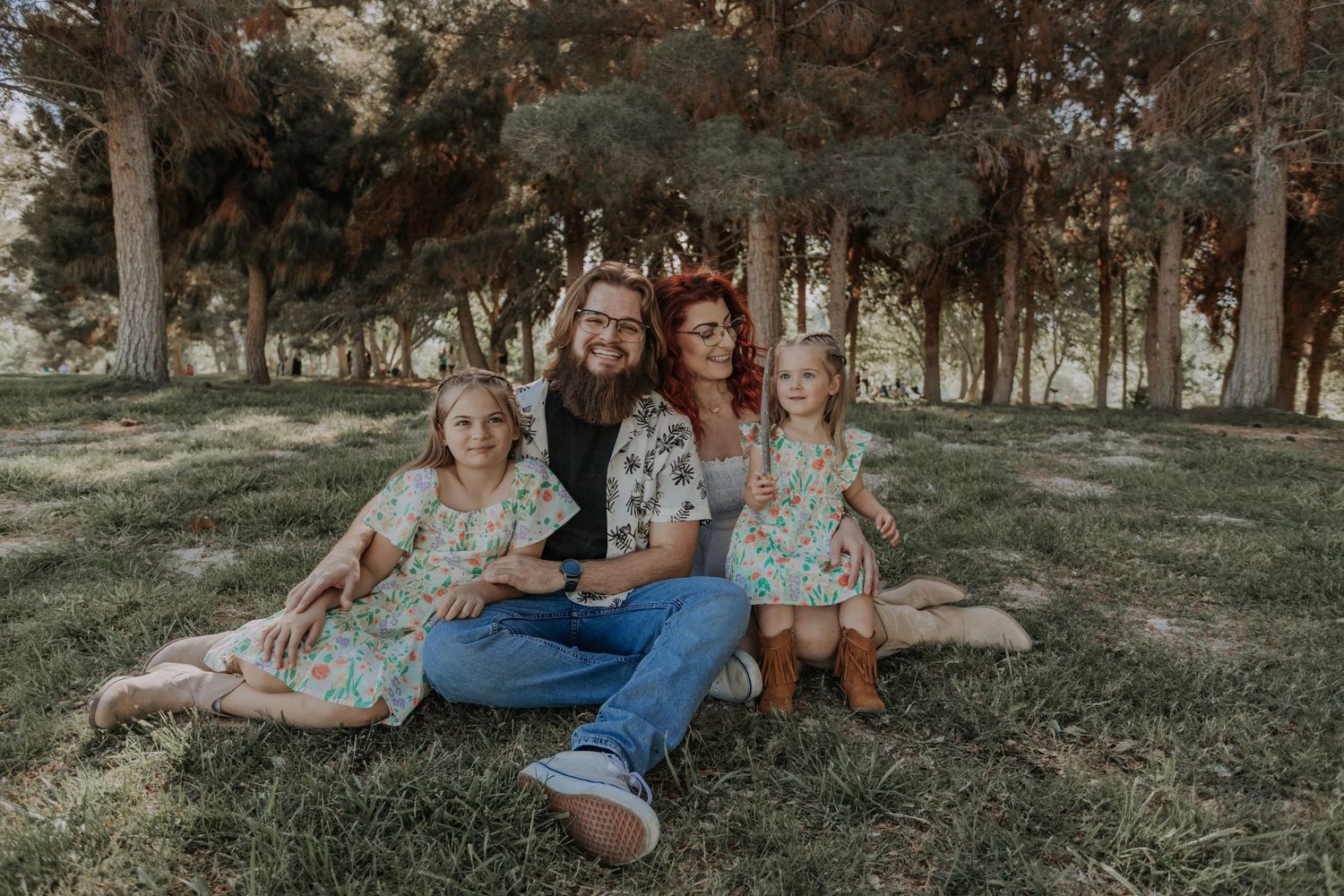 Family of four sitting on grass under pine trees, smiling together for a portrait.