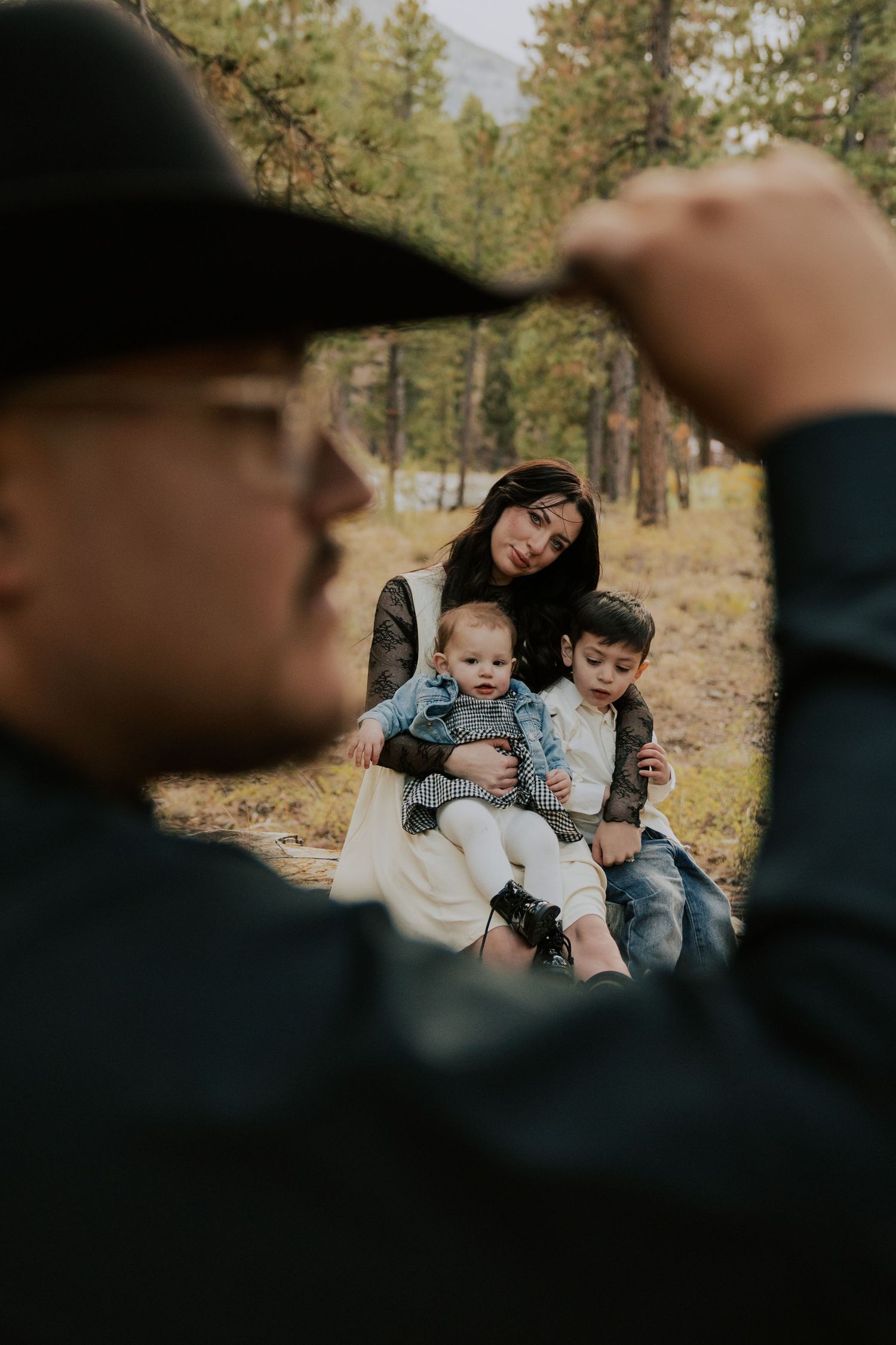 Mother sitting with two young children in a pine forest, framed by a man adjusting his hat.