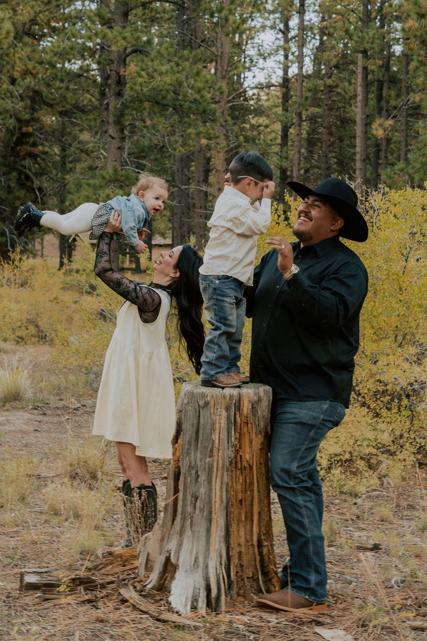 Family laughing and playing with two young children on a tree stump in a colorful autumn pine forest.